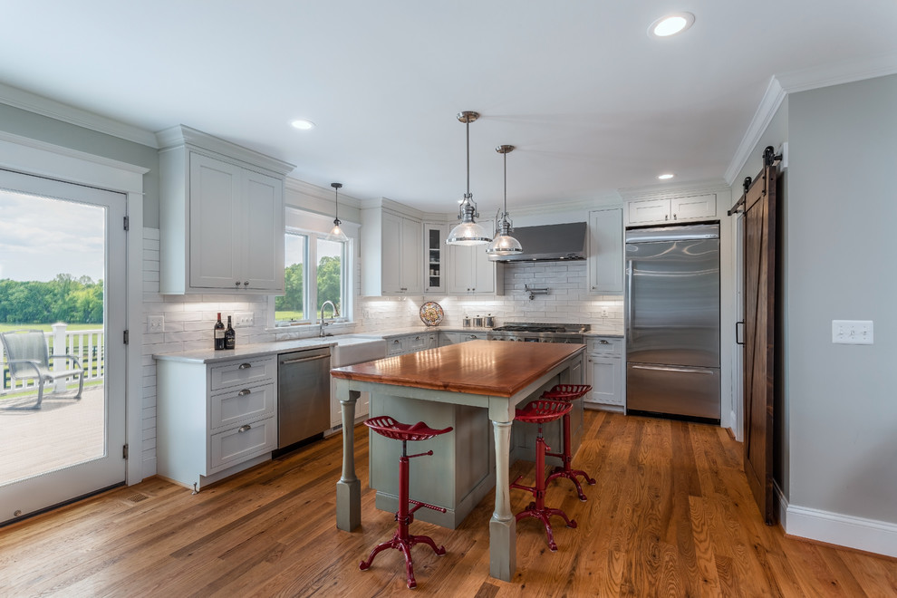 Design ideas for a country l-shaped open plan kitchen in DC Metro with a farmhouse sink, shaker cabinets, white cabinets, quartzite benchtops, white splashback, ceramic splashback, stainless steel appliances, light hardwood floors and with island.