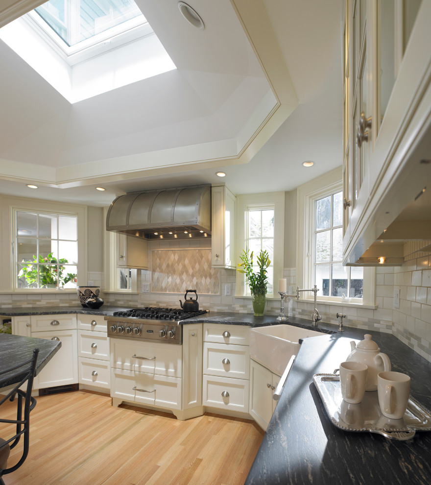 This is an example of a large country u-shaped eat-in kitchen in Boston with a farmhouse sink, recessed-panel cabinets, white cabinets, beige splashback, subway tile splashback, light hardwood floors, with island and stainless steel appliances.