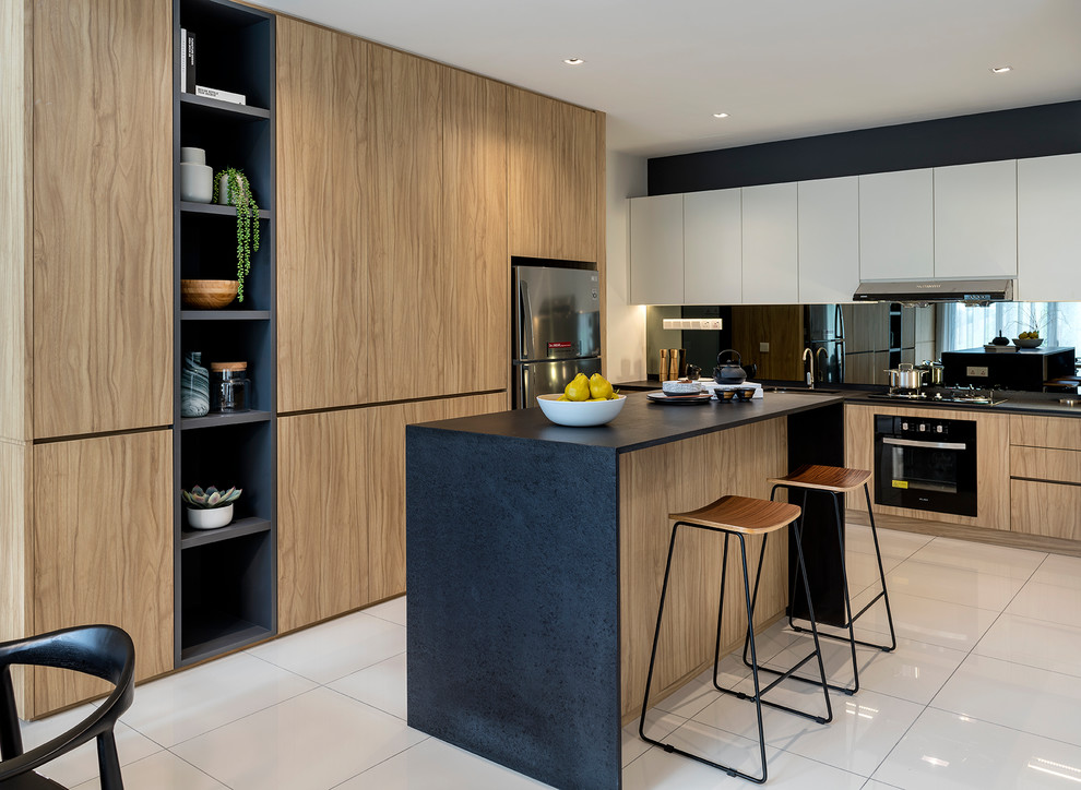This is an example of a contemporary l-shaped kitchen in Singapore with an undermount sink, flat-panel cabinets, medium wood cabinets, mirror splashback, with island, white floor and black benchtop.