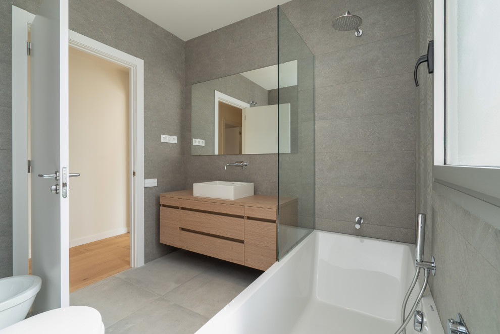 Photo of a mid-sized contemporary 3/4 bathroom in Barcelona with recessed-panel cabinets, brown cabinets, an alcove tub, a shower/bathtub combo, a wall-mount toilet, gray tile, ceramic tile, grey walls, ceramic floors, a vessel sink, wood benchtops, grey floor, an open shower, brown benchtops, an enclosed toilet, a single vanity, a built-in vanity, wallpaper and panelled walls.