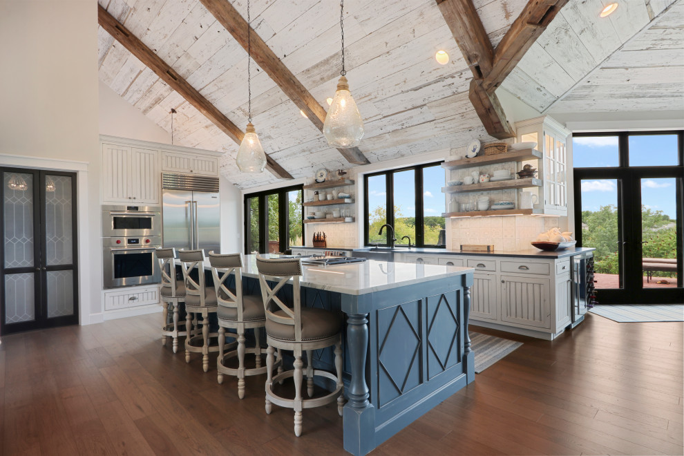 Country l-shaped open plan kitchen in Other with blue cabinets, ceramic splashback, stainless steel appliances, medium hardwood floors, multi-coloured benchtop, a farmhouse sink, white splashback, with island, brown floor and recessed-panel cabinets.