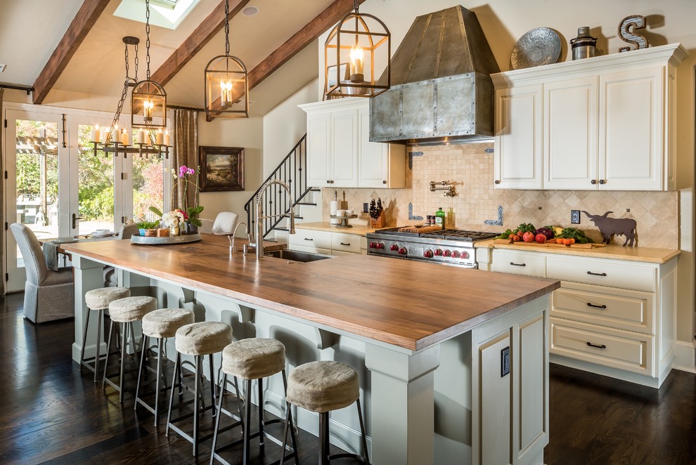 Example of a large transitional single-wall dark wood floor open concept kitchen design with a farmhouse sink, raised-panel cabinets, gray cabinets, wood countertops, beige backsplash, stone tile backsplash, stainless steel appliances and an island