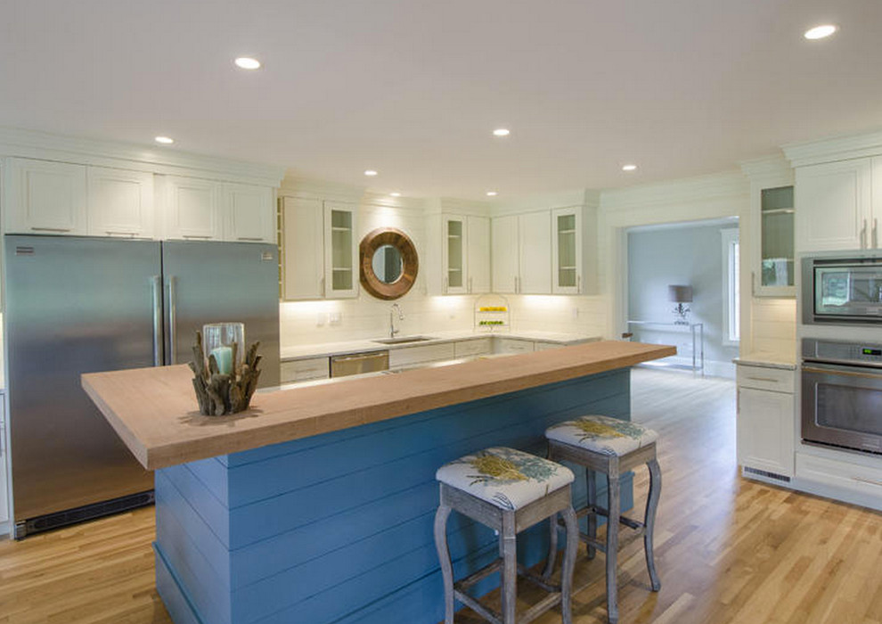 Large beach style l-shaped kitchen in Boston with a drop-in sink, glass-front cabinets, white cabinets, limestone benchtops, white splashback, stainless steel appliances, light hardwood floors and with island.