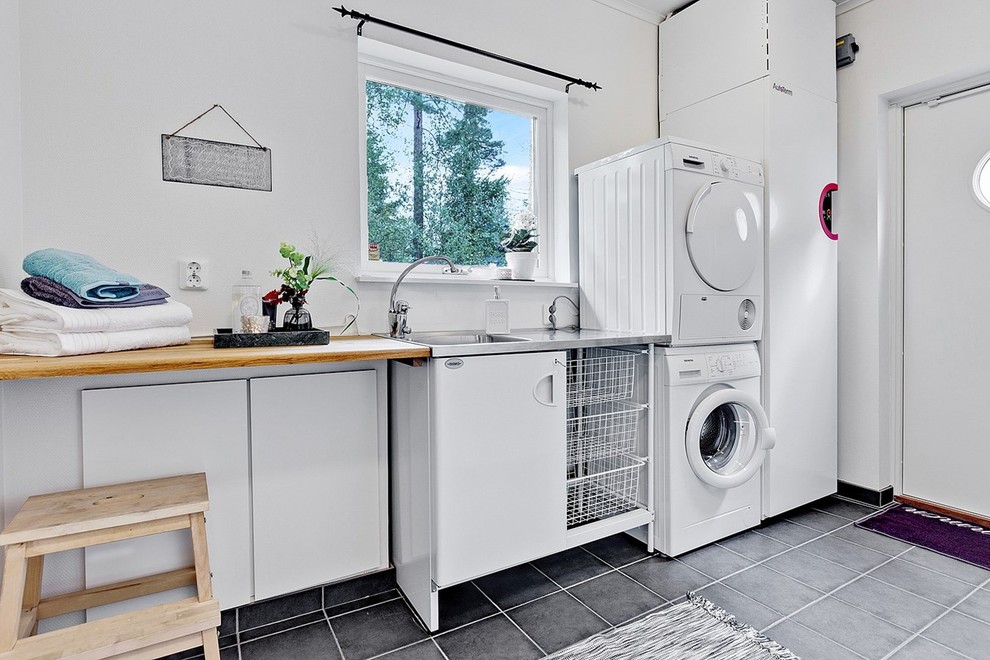 Design ideas for a mid-sized scandinavian single-wall dedicated laundry room in Stockholm with flat-panel cabinets, white cabinets, wood benchtops, white walls, a stacked washer and dryer, grey floor, beige benchtop and an integrated sink.