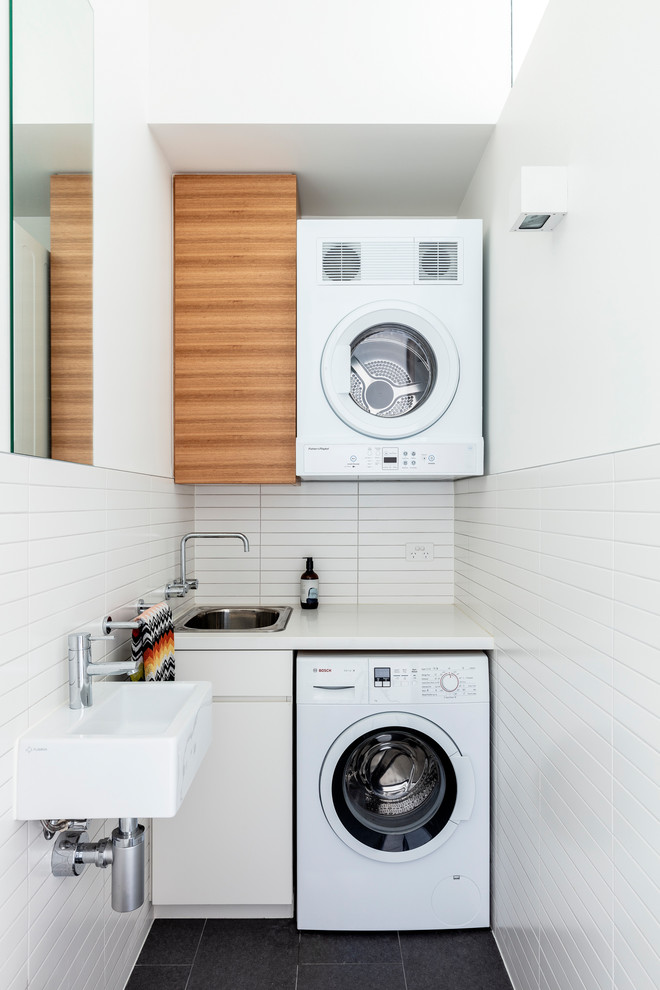 Inspiration for a mid-sized contemporary single-wall laundry room in Sydney with flat-panel cabinets, medium wood cabinets, white walls, a stacked washer and dryer, black floor and white benchtop.
