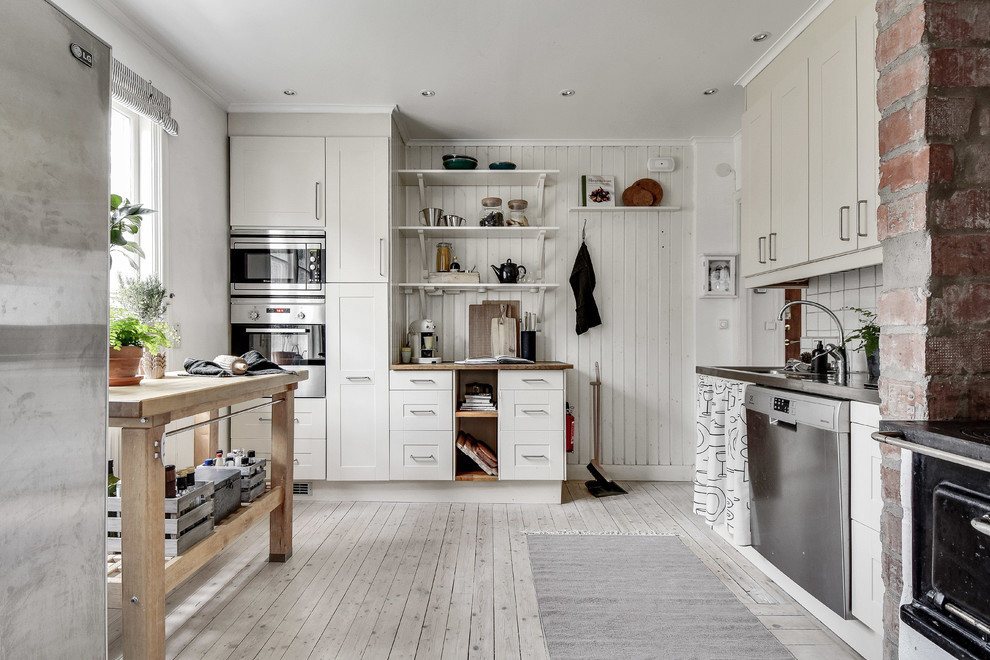 This is an example of a mid-sized country l-shaped separate kitchen in Stockholm with shaker cabinets, beige cabinets, wood benchtops, white splashback, stainless steel appliances, light hardwood floors and with island.