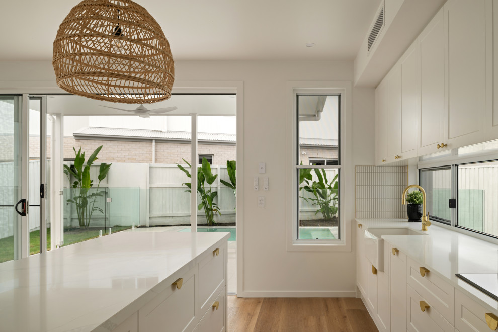 This is an example of a large tropical kitchen in Sunshine Coast with a farmhouse sink, shaker cabinets, white cabinets, marble benchtops, white splashback, matchstick tile splashback, black appliances, vinyl floors, with island and white benchtop.