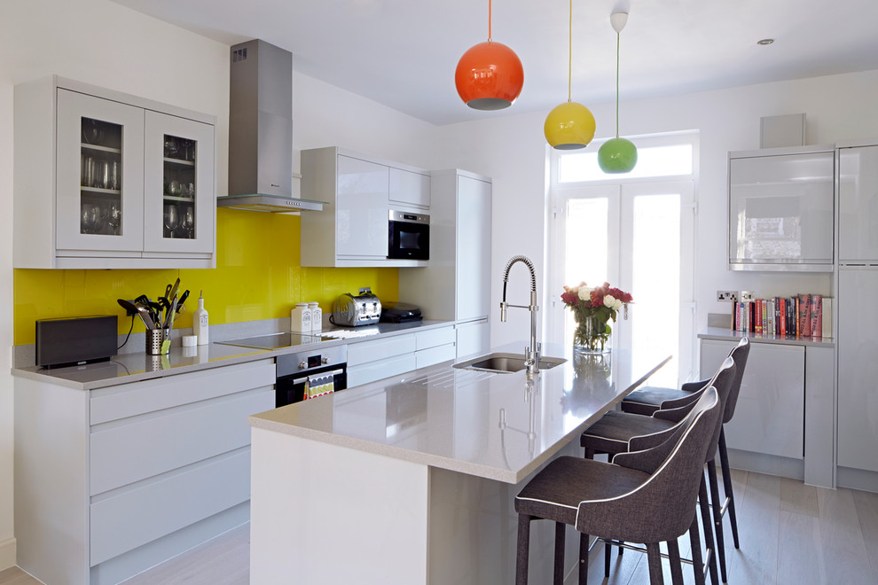 This is an example of a mid-sized contemporary galley kitchen in London with an undermount sink, flat-panel cabinets, white cabinets, quartz benchtops, yellow splashback, glass sheet splashback, light hardwood floors and with island.