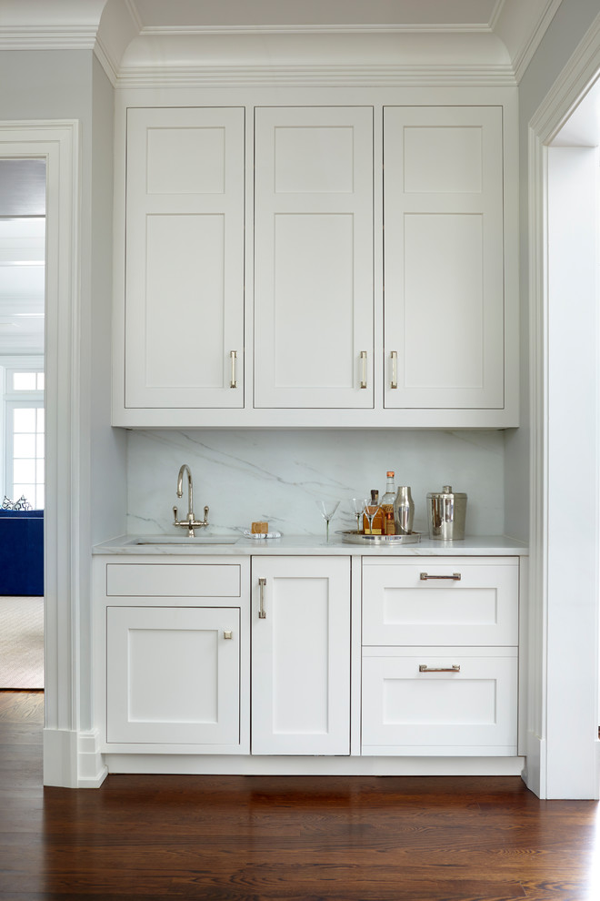 Photo of a small transitional single-wall wet bar in New York with shaker cabinets, white cabinets, marble benchtops, white splashback, marble splashback, medium hardwood floors, brown floor and white benchtop.