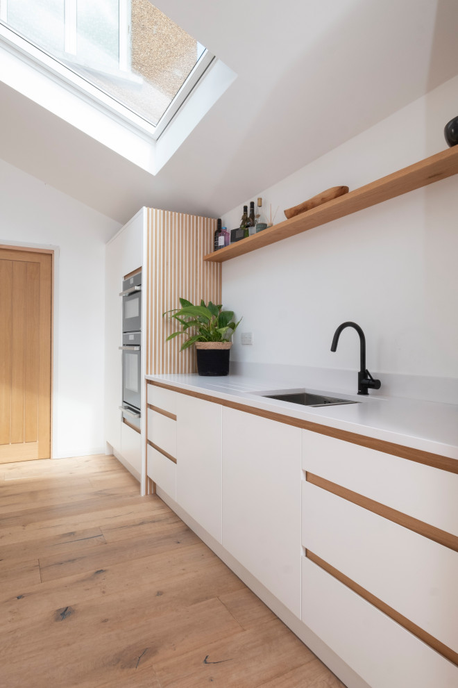 Design ideas for a mid-sized scandinavian single-wall open plan kitchen in Cardiff with an integrated sink, flat-panel cabinets, white cabinets, quartzite benchtops, white splashback, stainless steel appliances, light hardwood floors, with island, white benchtop and vaulted.