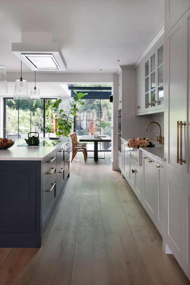 Inspiration for a large single-wall eat-in kitchen in London with a farmhouse sink, raised-panel cabinets, white cabinets, solid surface benchtops, white splashback, ceramic splashback, stainless steel appliances, light hardwood floors, with island, brown floor and white benchtop.