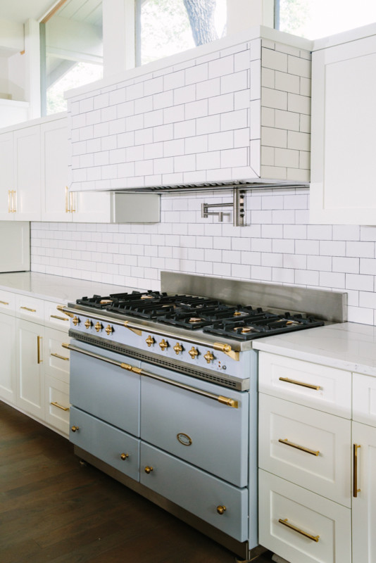 Southill - We completely gutted the client's existing kitchen and opened up the space.  We kept the windows bare to let in beautiful light and showcase the oak trees outside, used oversized unlacquered brass cabinet hardware, and painted the island a dark to contrast against the light tile, cabinetry and countertops. We brought in a pop of subtle color with a Lacanche range imported from France.