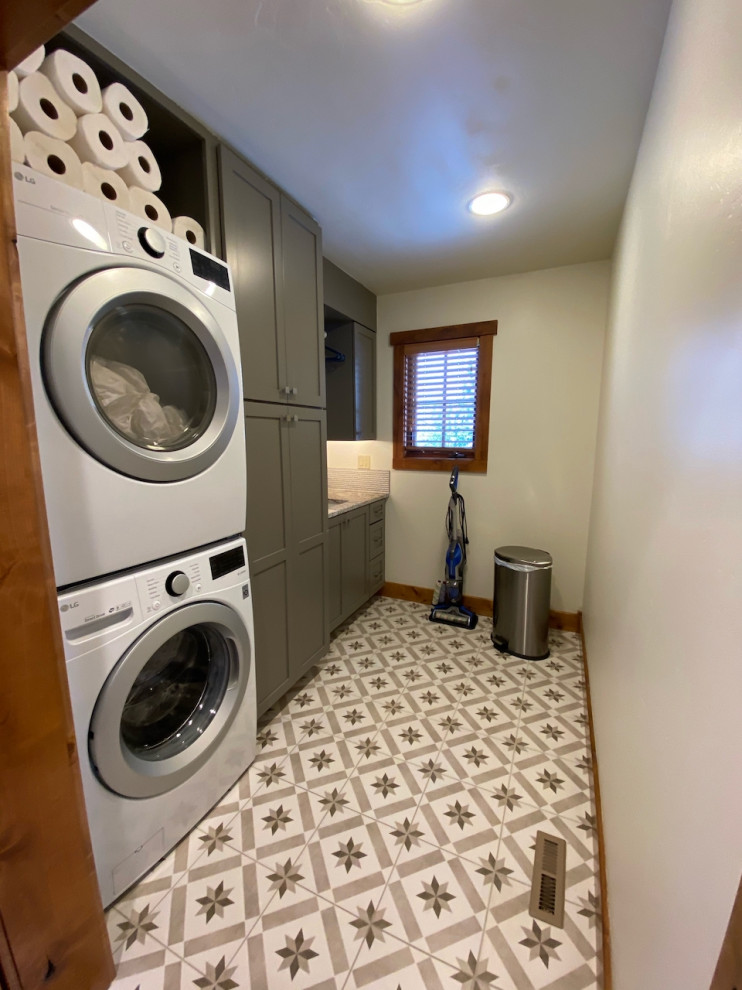 This is an example of a mid-sized transitional galley dedicated laundry room in Boise with an undermount sink, shaker cabinets, grey cabinets, granite benchtops, white splashback, mosaic tile splashback, white walls, porcelain floors, a stacked washer and dryer, beige floor and multi-coloured benchtop.