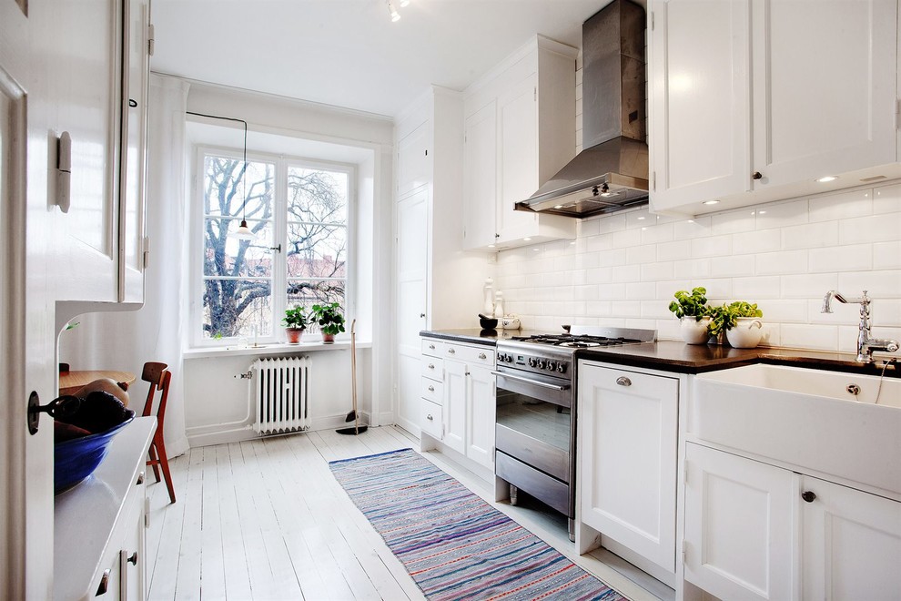 Design ideas for a scandinavian galley eat-in kitchen in Stockholm with a farmhouse sink, shaker cabinets, white cabinets, white splashback, subway tile splashback, stainless steel appliances, painted wood floors, wood benchtops and no island.