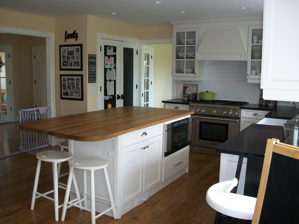 This is an example of a mid-sized country l-shaped eat-in kitchen in Calgary with a farmhouse sink, shaker cabinets, white cabinets, wood benchtops, white splashback, porcelain splashback, stainless steel appliances, light hardwood floors and with island.