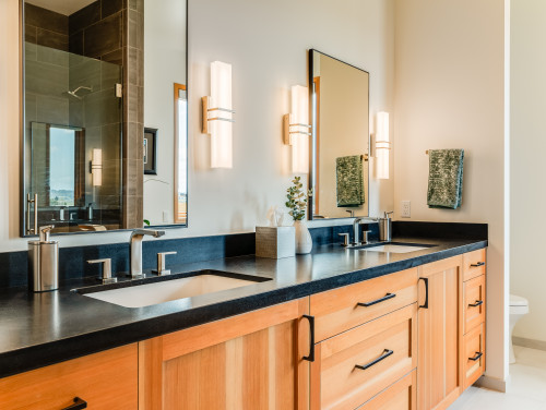 Natural wood cabinets with matte black pulls, black quartz countertop, dual sinks, chrome faucets, frameless mirrors, and modern wall sconces