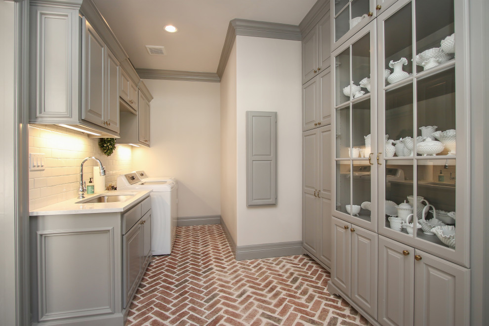 Inspiration for a large traditional u-shaped utility room in Houston with an undermount sink, raised-panel cabinets, grey cabinets, white splashback, subway tile splashback, grey walls, brick floors, a side-by-side washer and dryer, red floor and white benchtop.
