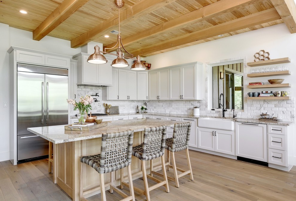 Inspiration for a country l-shaped kitchen in Austin with a farmhouse sink, shaker cabinets, white cabinets, white splashback, stainless steel appliances, light hardwood floors, with island, beige floor and grey benchtop.