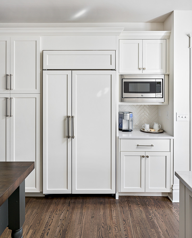 This is an example of a mid-sized traditional u-shaped eat-in kitchen in Chicago with a farmhouse sink, shaker cabinets, white cabinets, quartz benchtops, white splashback, porcelain splashback, stainless steel appliances, dark hardwood floors, a peninsula and brown floor.