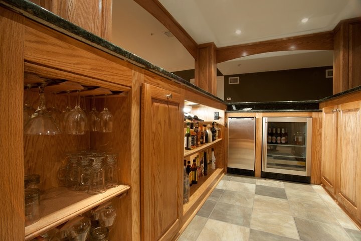 This is an example of a large traditional u-shaped seated home bar in Philadelphia with an undermount sink, raised-panel cabinets, medium wood cabinets, granite benchtops, porcelain floors, grey floor and black benchtop.