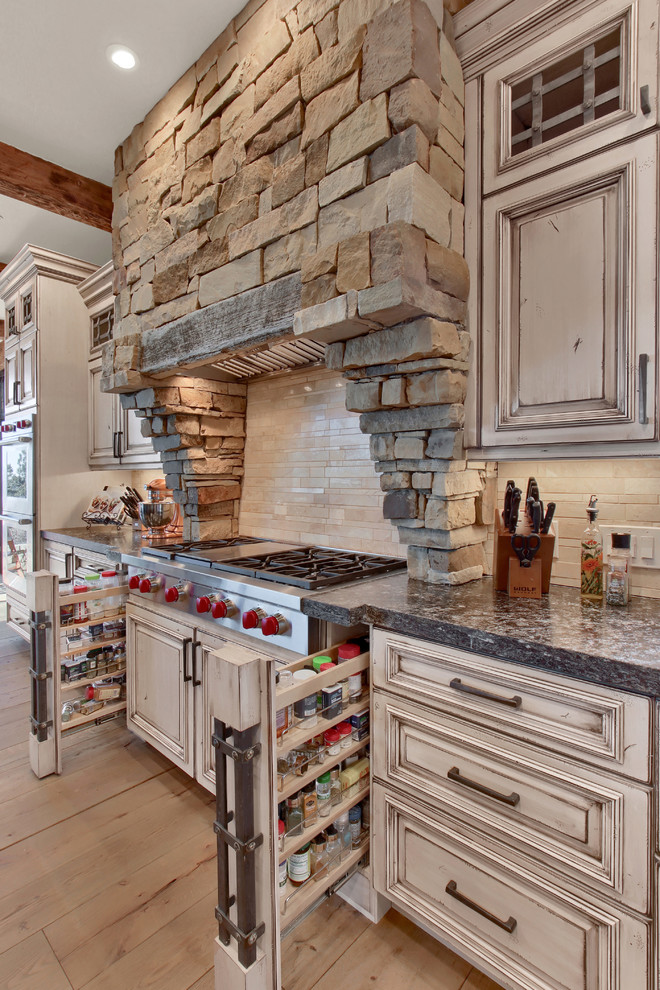 This is an example of a large country l-shaped open plan kitchen in Denver with a farmhouse sink, raised-panel cabinets, white cabinets, granite benchtops, beige splashback, travertine splashback, panelled appliances, light hardwood floors, with island, beige floor and grey benchtop.