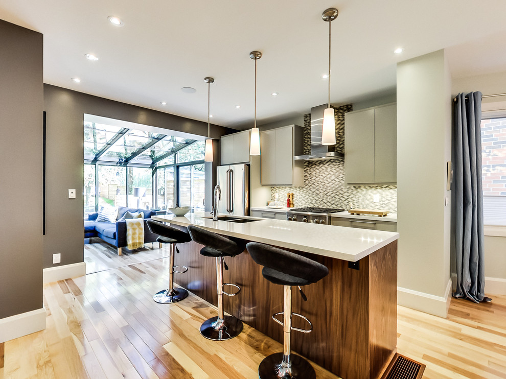 Inspiration for a mid-sized transitional galley light wood floor and multicolored floor kitchen remodel in Toronto with a double-bowl sink, flat-panel cabinets, beige cabinets, quartz countertops, gray backsplash, ceramic backsplash, stainless steel appliances and an island