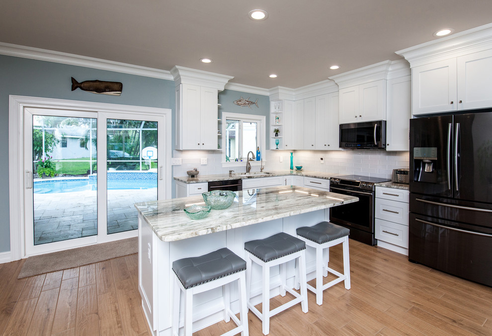 Photo of a mid-sized beach style l-shaped open plan kitchen in Tampa with shaker cabinets, white cabinets, granite benchtops, white splashback, subway tile splashback, stainless steel appliances, light hardwood floors, with island, brown floor and beige benchtop.