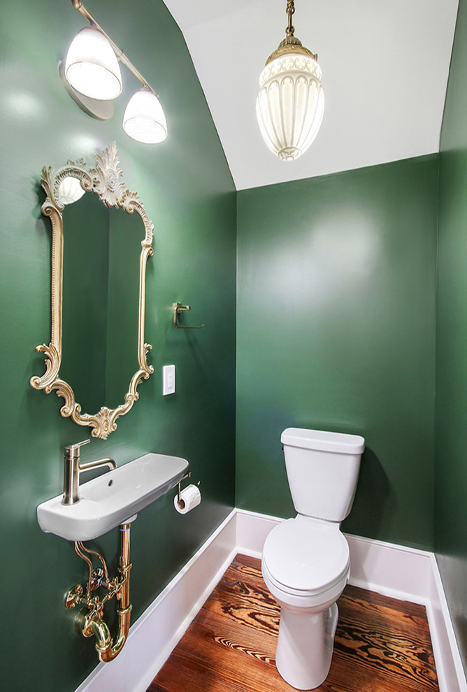 This is an example of a small traditional powder room with green walls, medium hardwood floors, a wall-mount sink, brown floor and a two-piece toilet.