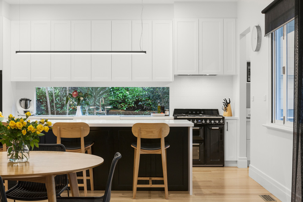 Mid-sized contemporary single-wall open plan kitchen in Melbourne with a farmhouse sink, shaker cabinets, white cabinets, quartz benchtops, white splashback, window splashback, black appliances, light hardwood floors, with island, beige floor and white benchtop.