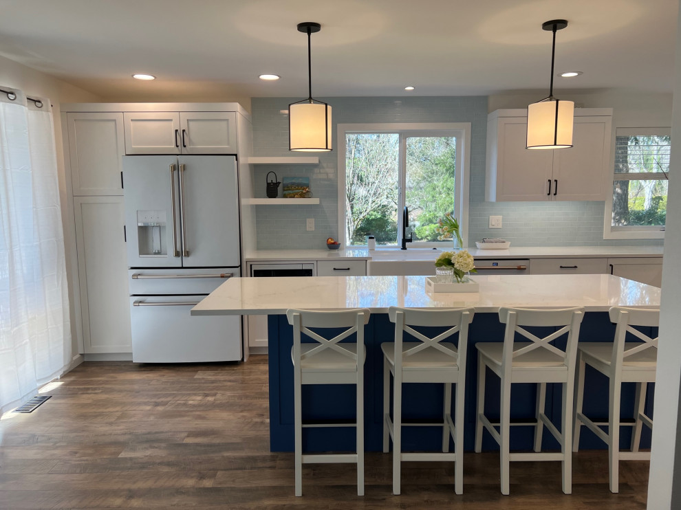 Foto på ett mellanstort vintage vit kök, med en rustik diskho, skåp i shakerstil, blå skåp, bänkskiva i kvarts, blått stänkskydd, stänkskydd i keramik, vita vitvaror, laminatgolv, en köksö och brunt golv