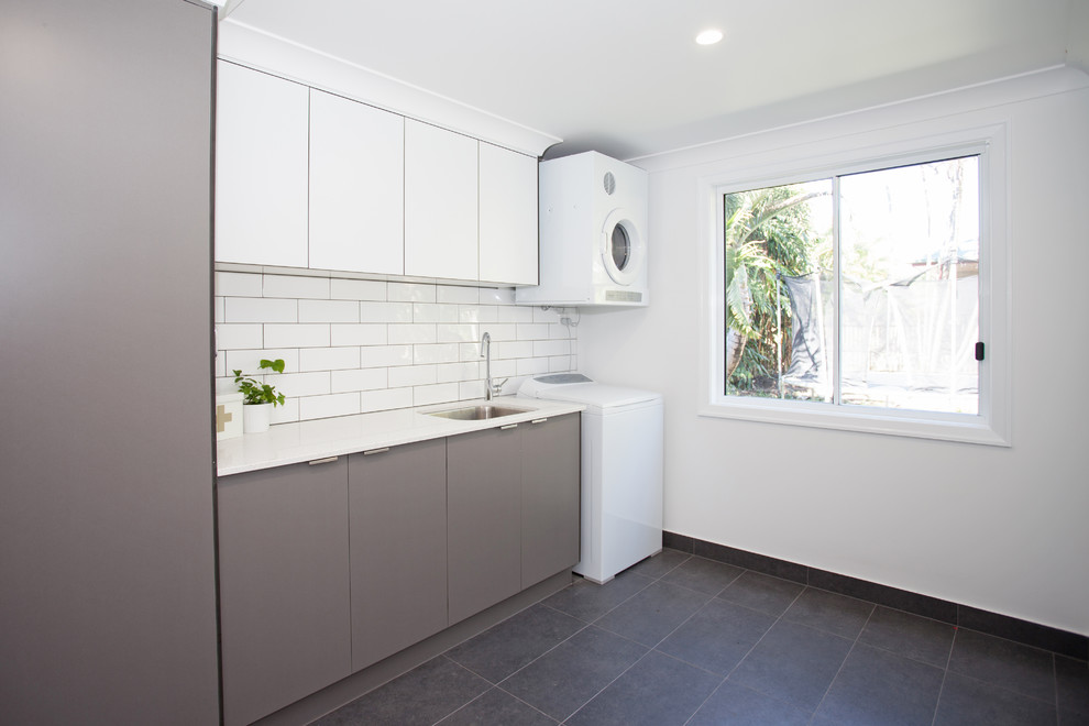 Inspiration for a large tropical single-wall dedicated laundry room with an undermount sink, flat-panel cabinets, grey cabinets, quartz benchtops, white walls, a stacked washer and dryer and black floor.