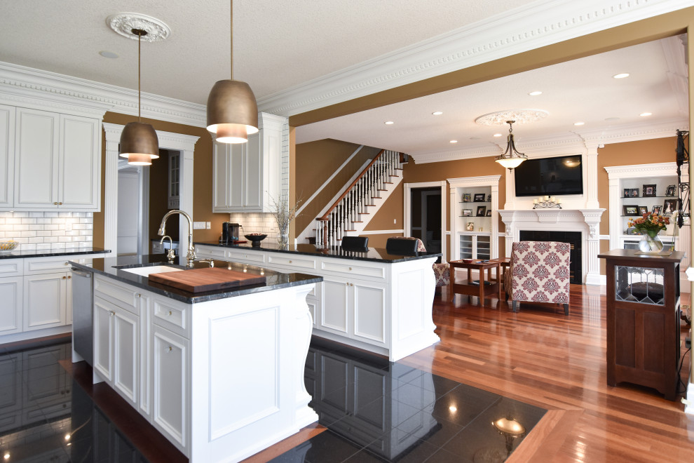 This is an example of a traditional u-shaped open plan kitchen in Edmonton with an undermount sink, shaker cabinets, white cabinets, granite benchtops, white splashback, subway tile splashback, stainless steel appliances, medium hardwood floors, multiple islands, brown floor and black benchtop.
