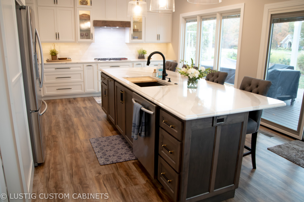 Open concept kitchen - large transitional l-shaped laminate floor and brown floor open concept kitchen idea with an undermount sink, flat-panel cabinets, white cabinets, quartz countertops, white backsplash, ceramic backsplash, stainless steel appliances, an island and white countertops