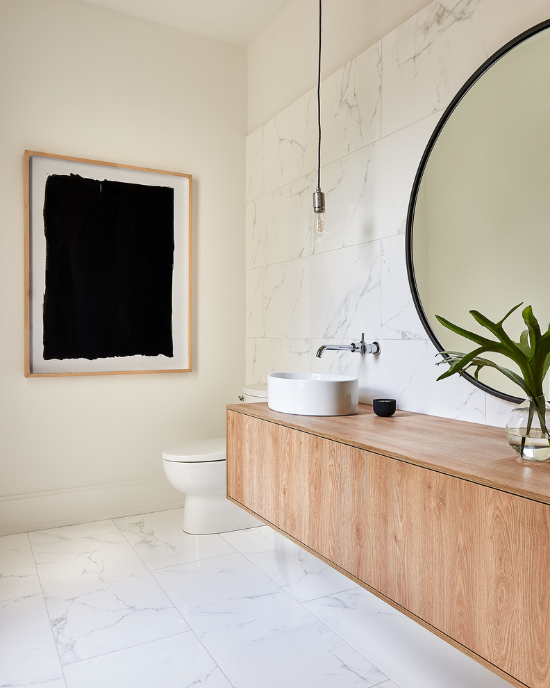 Inspiration for a large contemporary powder room in Charlotte with flat-panel cabinets, light wood cabinets, white tile, marble, white walls, marble floors, a vessel sink, wood benchtops, white floor and beige benchtops.
