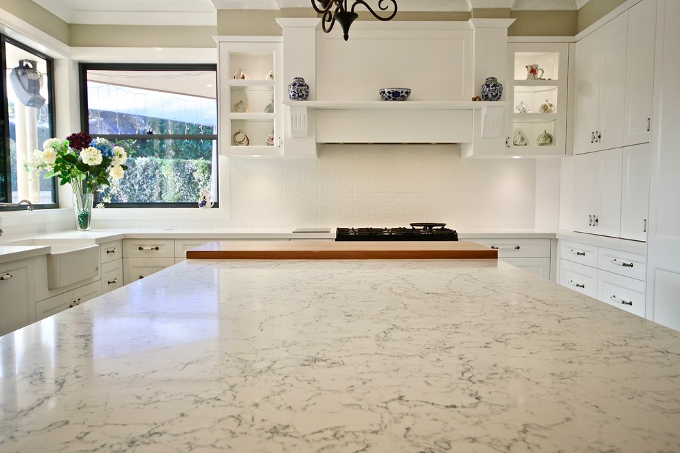 This is an example of a mid-sized transitional u-shaped eat-in kitchen in Sydney with a farmhouse sink, shaker cabinets, white cabinets, quartz benchtops, white splashback, porcelain splashback, stainless steel appliances, porcelain floors, with island and beige floor.