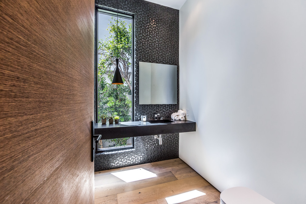 Mid-sized modern powder room in Los Angeles with a one-piece toilet, black tile, light hardwood floors, an integrated sink, white walls and black benchtops.