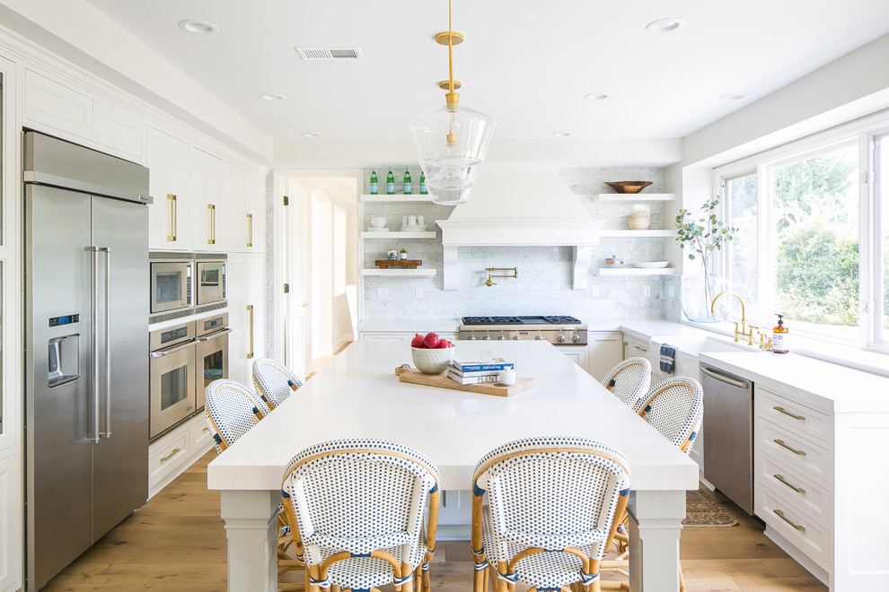 This is an example of a beach style u-shaped kitchen in Los Angeles with a farmhouse sink, recessed-panel cabinets, white cabinets, stainless steel appliances, medium hardwood floors, with island and brown floor.