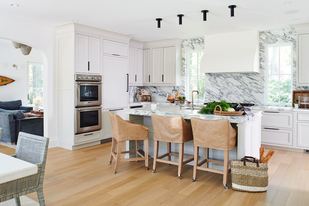 Inspiration for a beach style l-shaped kitchen in New York with an undermount sink, shaker cabinets, white cabinets, white splashback, stone slab splashback, stainless steel appliances, light hardwood floors, with island, beige floor and white benchtop.
