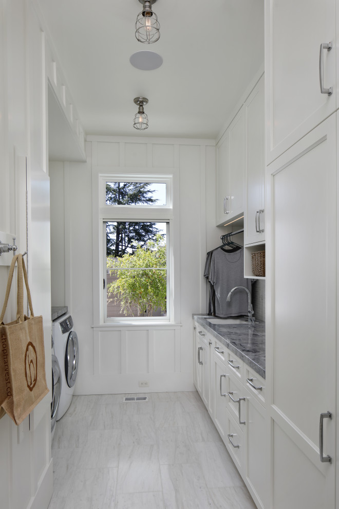 Idéer för lantliga parallella grått tvättstugor enbart för tvätt, med en undermonterad diskho, luckor med infälld panel, vita skåp, marmorbänkskiva, vitt stänkskydd, stänkskydd i glaskakel, vita väggar, klinkergolv i keramik, en tvättmaskin och torktumlare bredvid varandra och vitt golv