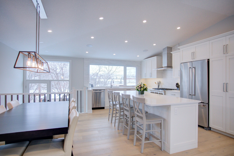 Mid-sized contemporary l-shaped open plan kitchen in Calgary with an undermount sink, shaker cabinets, white cabinets, quartz benchtops, white splashback, ceramic splashback, stainless steel appliances, light hardwood floors, with island, beige floor and white benchtop.