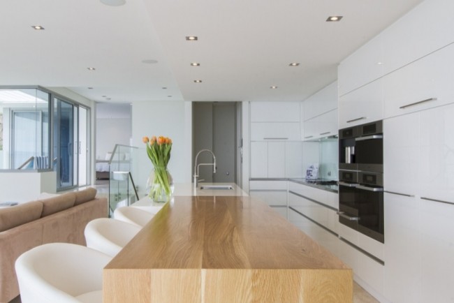 This is an example of a modern galley eat-in kitchen in Perth with an integrated sink, flat-panel cabinets, white cabinets, wood benchtops, white splashback, glass sheet splashback, stainless steel appliances, travertine floors and with island.