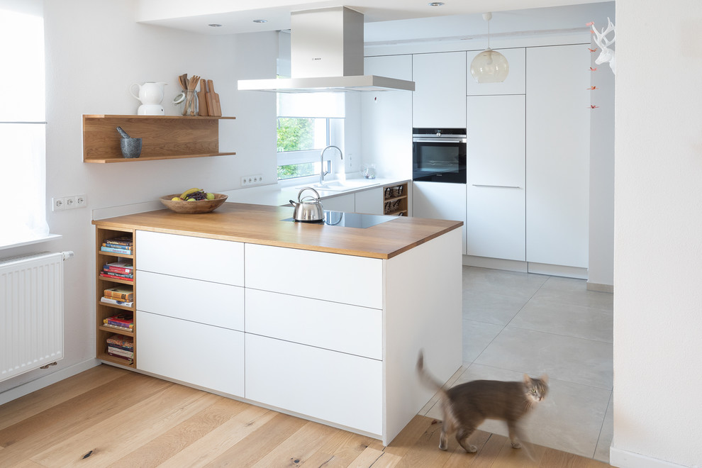 Inspiration for a mid-sized contemporary u-shaped open plan kitchen in Stuttgart with a drop-in sink, flat-panel cabinets, white cabinets, wood benchtops, white splashback, black appliances, limestone floors, beige floor, brown benchtop and a peninsula.