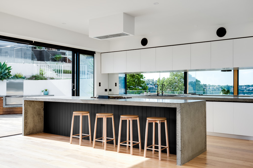 This is an example of a contemporary galley kitchen in Sydney with white cabinets, concrete benchtops, window splashback, medium hardwood floors, with island, brown floor and grey benchtop.