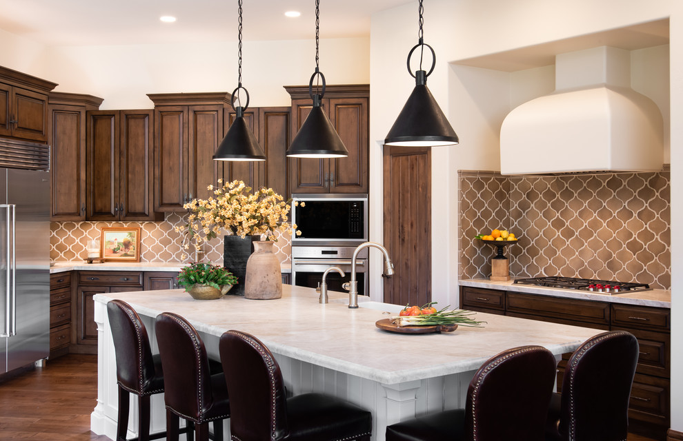 This is an example of a mid-sized mediterranean l-shaped eat-in kitchen in Phoenix with a farmhouse sink, raised-panel cabinets, quartzite benchtops, terra-cotta splashback, stainless steel appliances, medium hardwood floors, with island, brown floor, beige benchtop and brown cabinets.