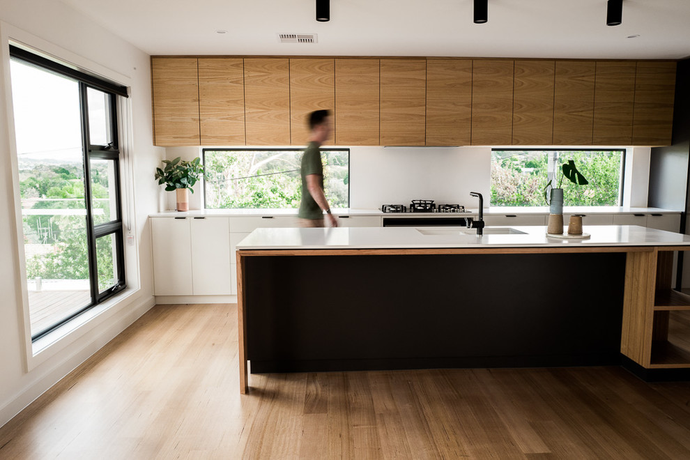 This is an example of a contemporary galley kitchen in Canberra - Queanbeyan with an undermount sink, quartz benchtops, white splashback, stone slab splashback, black appliances, light hardwood floors and white benchtop.