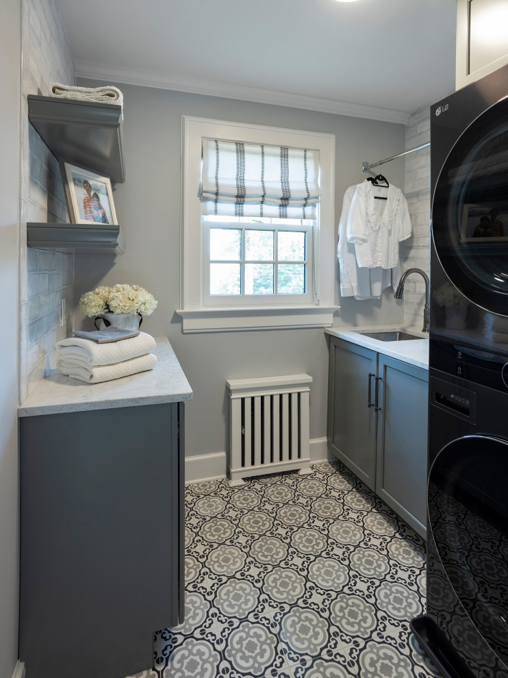 This is an example of a mid-sized traditional u-shaped dedicated laundry room in New York with an undermount sink, shaker cabinets, green cabinets, solid surface benchtops, white splashback, ceramic splashback, grey walls, concrete floors, a stacked washer and dryer, grey floor and white benchtop.