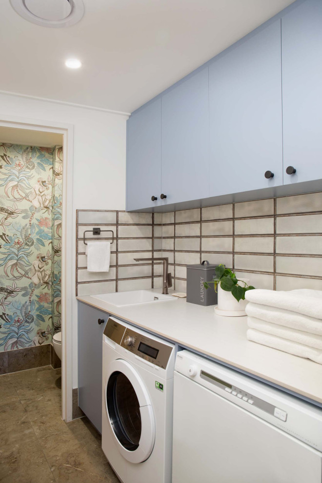 This is an example of a small industrial single-wall utility room in Brisbane with a belfast sink, flat-panel cabinets, blue cabinets, composite countertops, grey splashback, ceramic splashback, multi-coloured walls, ceramic flooring, a side by side washer and dryer, brown floors, white worktops and wallpapered walls.