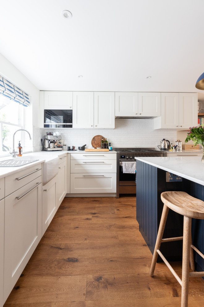 Large kitchen in London with a belfast sink, shaker cabinets, white cabinets, white splashback, stainless steel appliances, medium hardwood flooring, an island, brown floors and feature lighting.