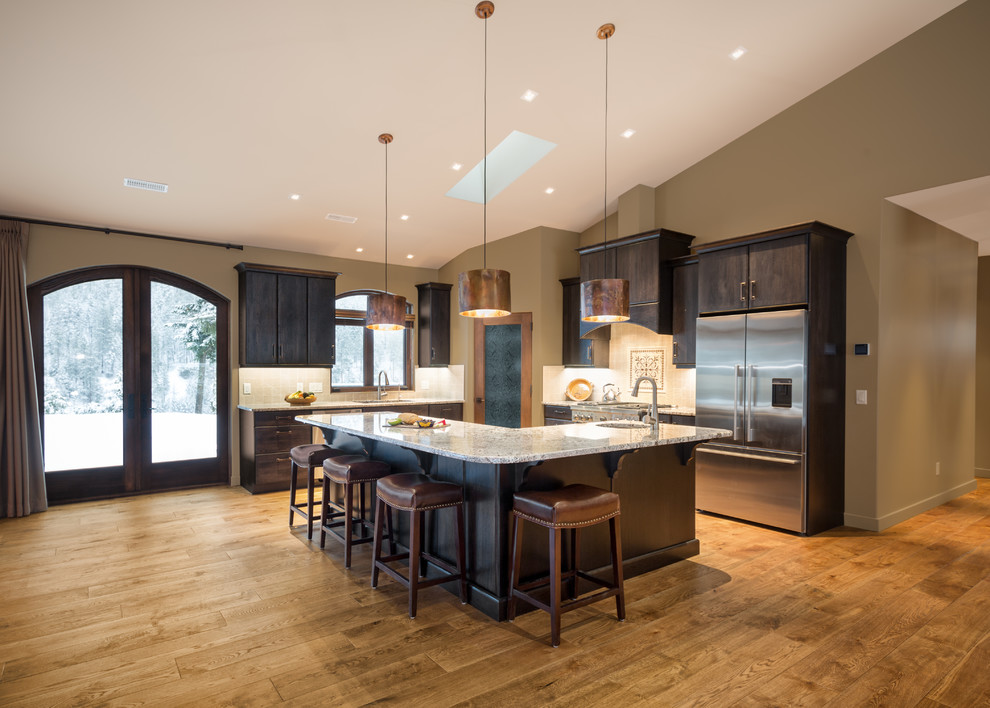 Design ideas for a large country l-shaped open plan kitchen in Vancouver with flat-panel cabinets, dark wood cabinets, with island, an undermount sink, granite benchtops, beige splashback, stone tile splashback, stainless steel appliances and medium hardwood floors.