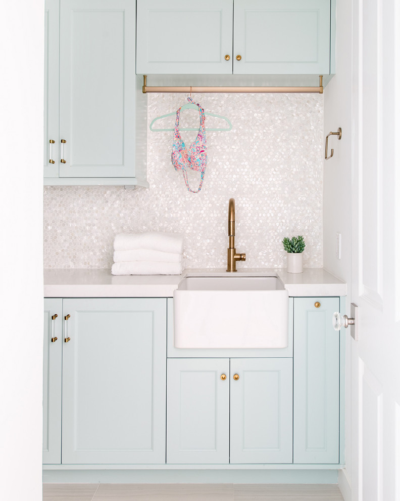 Inspiration for a small beach style dedicated laundry room in Los Angeles with a farmhouse sink, recessed-panel cabinets, quartzite benchtops, white splashback, white walls, white benchtop, blue cabinets, mosaic tile splashback and grey floor.